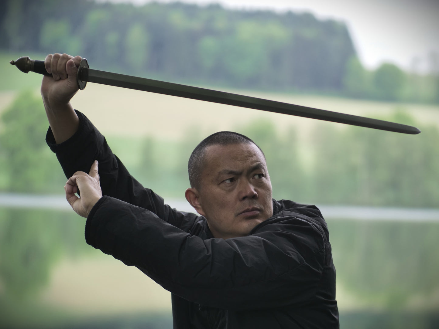 Person in black clothing holding a sword outdoors with greenery in the background