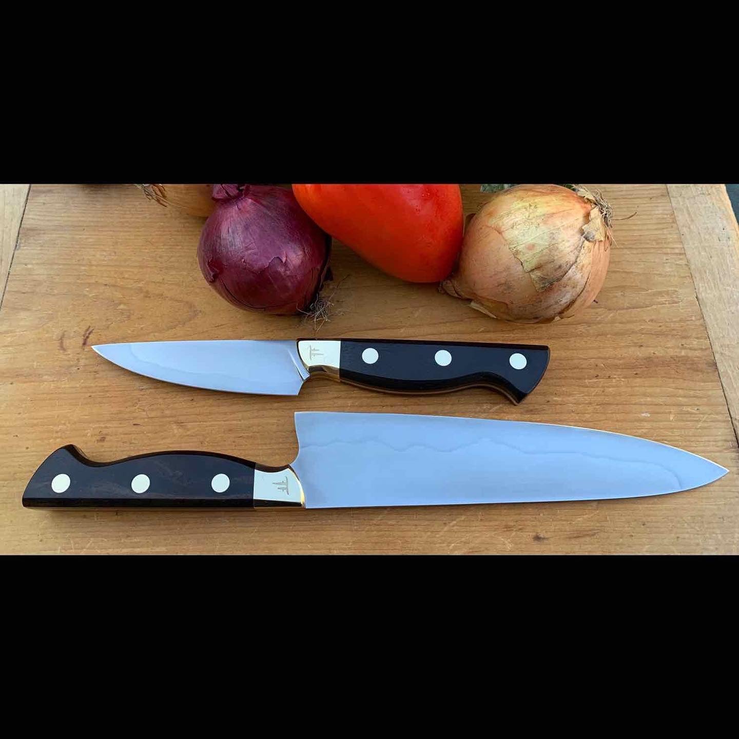 Two chef's knives with Hamon on a wooden cutting board with vegetables in the background.