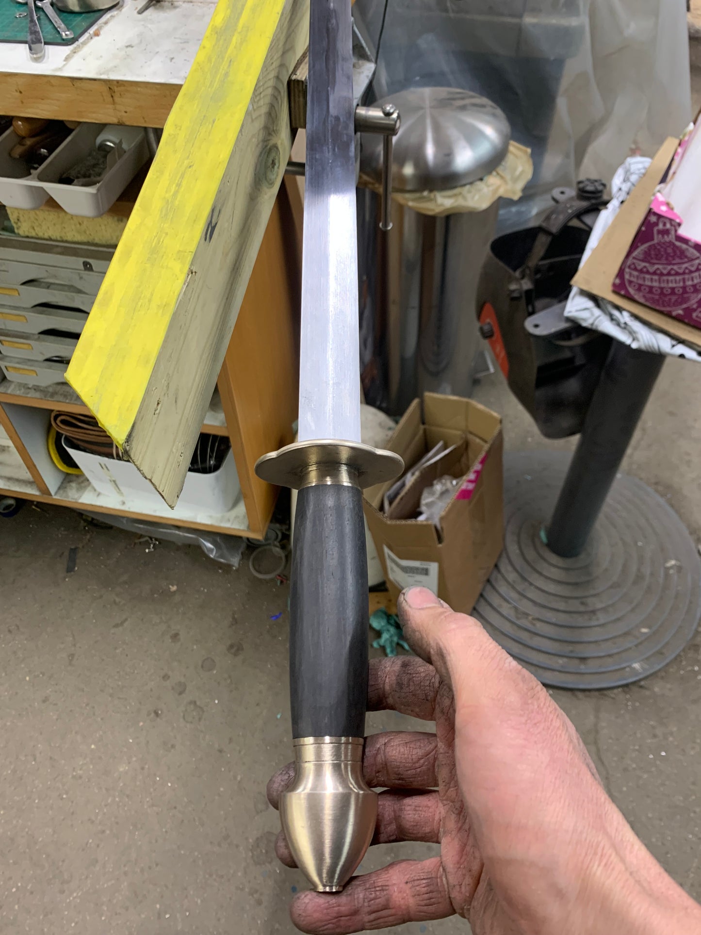 Hand holding a sword with a black and gold handle in a workshop setting