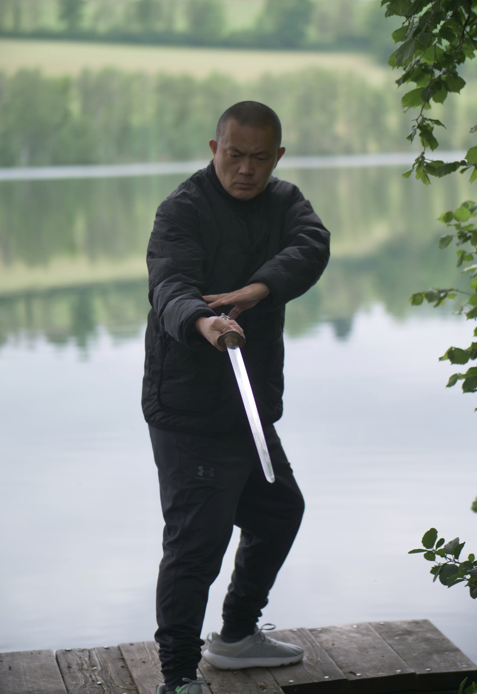 Person holding a sword by a body of water with trees in the background